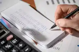 A hand recording financial figures in a notebook beside a calculator